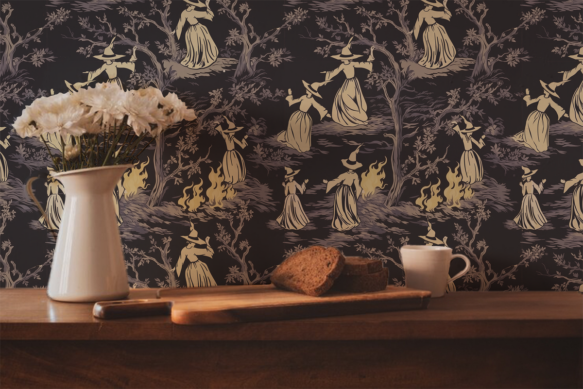 Moody peel-and-stick wallpaper sample featuring a witchy toile pattern with cream-cloaked figures, twisted gray trees, and glowing bonfire details on a deep charcoal background.