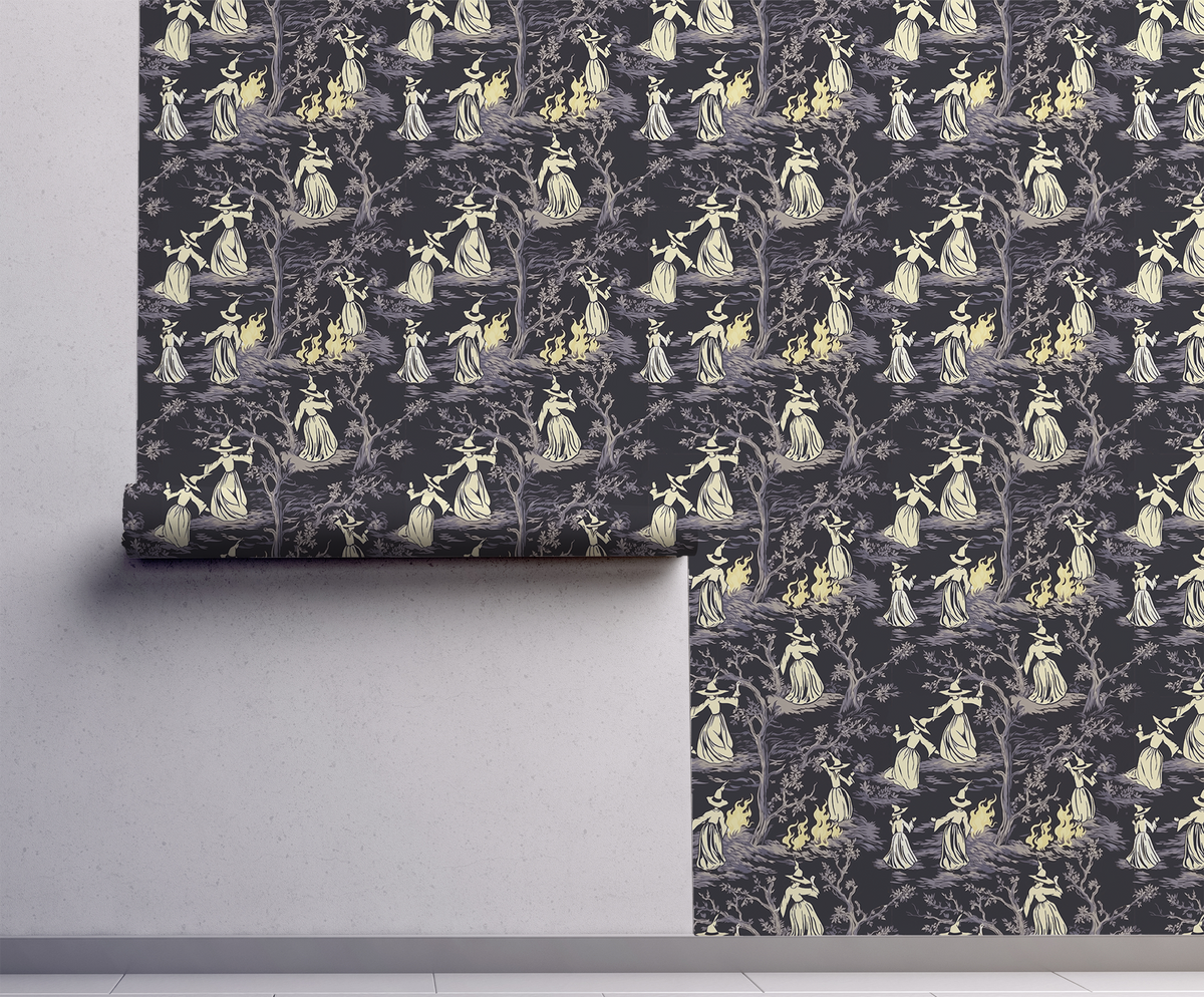 Moody peel-and-stick wallpaper sample featuring a witchy toile pattern with cream-cloaked figures, twisted gray trees, and glowing bonfire details on a deep charcoal background on wall