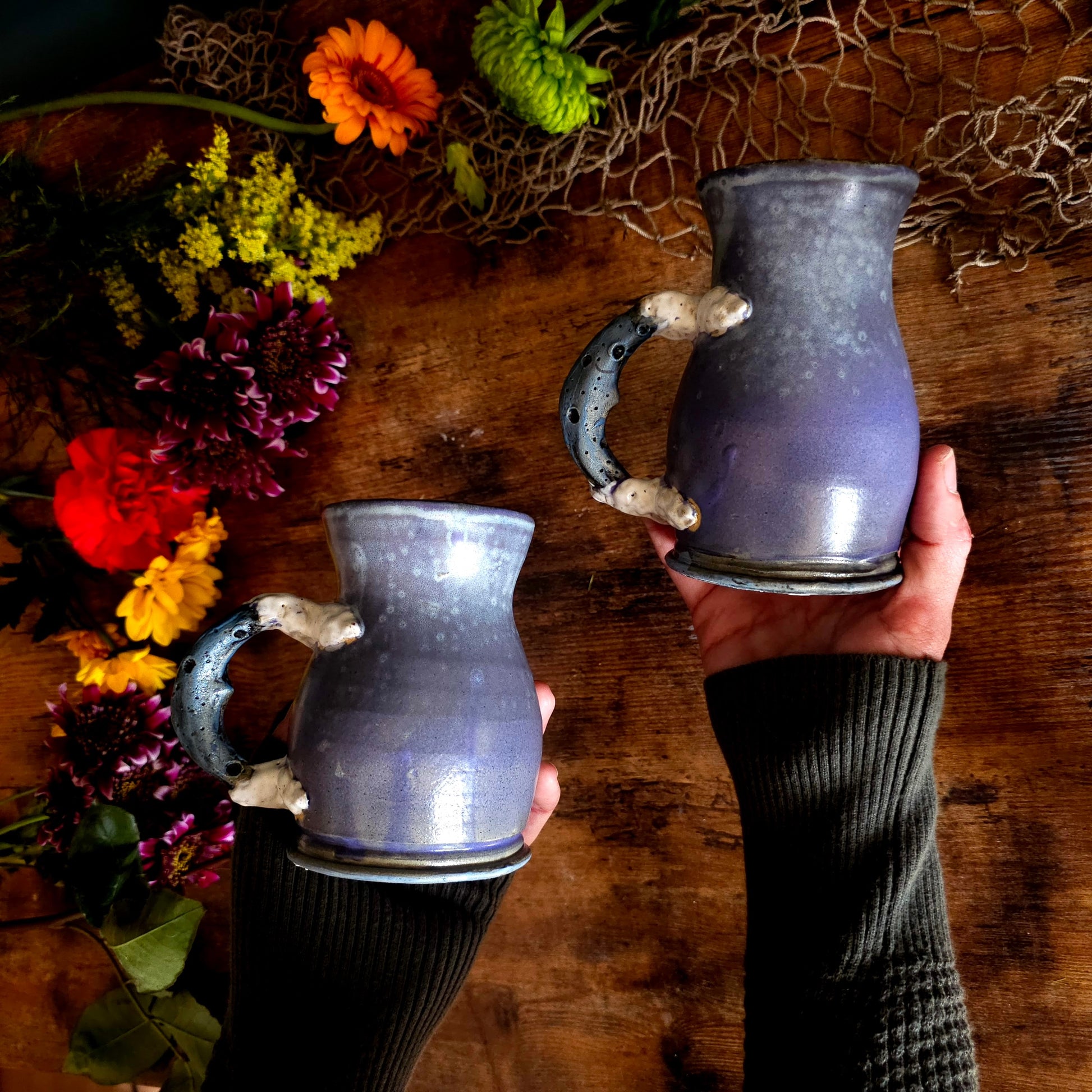 Handmade lavender ceramic mug with moon crater handle on rustic wood table, moody cottagecore backdrop with dark green wall and floral jar.