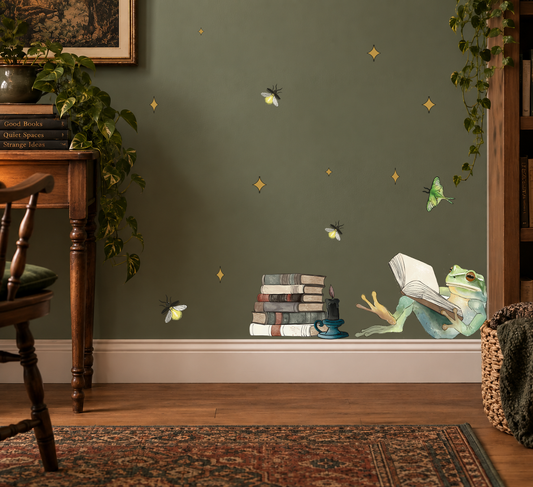 A styled room scene with a dark green wall decorated with whimsical wall decals, including a green frog reading a book, a stack of books, fireflies, and small gold stars. The room features a wooden table, a potted plant, a patterned rug, and a bookshelf.