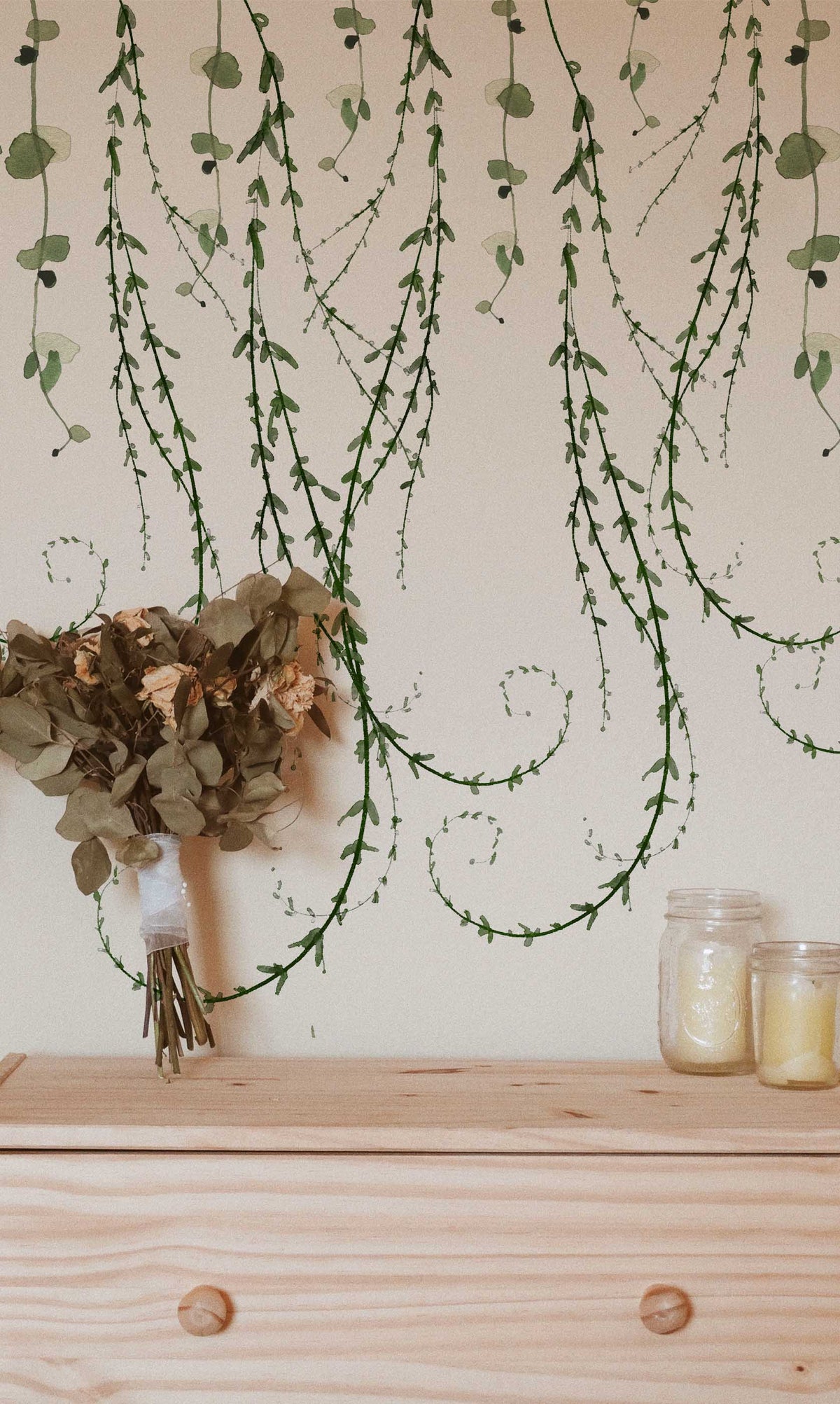 Trailing Vines botanical peel and stick wallpaper with cascading green vines on a cream wall in a cottage-style bedroom.