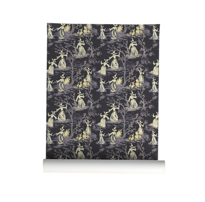 Moody peel-and-stick wallpaper sample featuring a witchy toile pattern with cream-cloaked figures, twisted gray trees, and glowing bonfire details on a deep charcoal background on roll