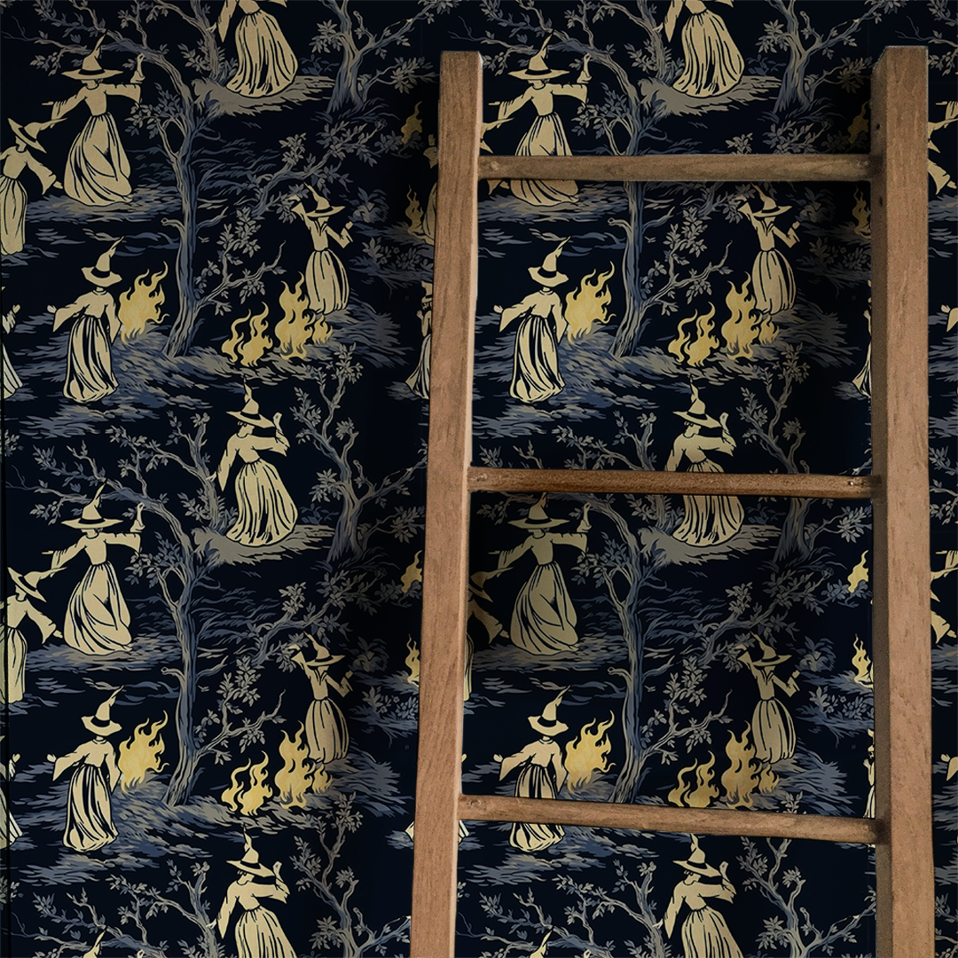 Moody peel-and-stick wallpaper sample featuring a witchy toile pattern with cream-cloaked figures, twisted gray trees, and glowing bonfire details on a deep charcoal background on wall