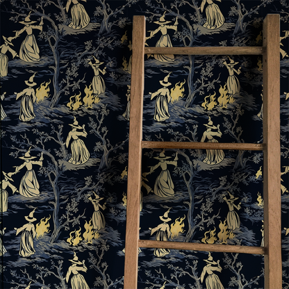 Moody peel-and-stick wallpaper sample featuring a witchy toile pattern with cream-cloaked figures, twisted gray trees, and glowing bonfire details on a deep charcoal background on wall