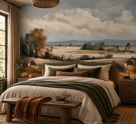 A styled bedroom featuring a large wall mural of a pastoral landscape with rolling fields, trees, and a cloudy sky. The bed is dressed in cream bedding with green and rust accent pillows and throws, and the room is lit by warm bedside lamps and natural light from a window.